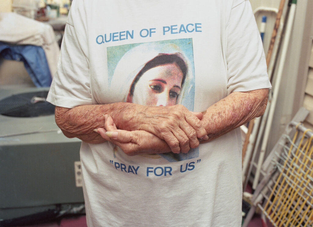 Woman’s arm crossed over a t-shirt with the image of the Virgin Mary that reads ‘QUEEN OF PEACE “PRAY FOR US”’ capital letters.