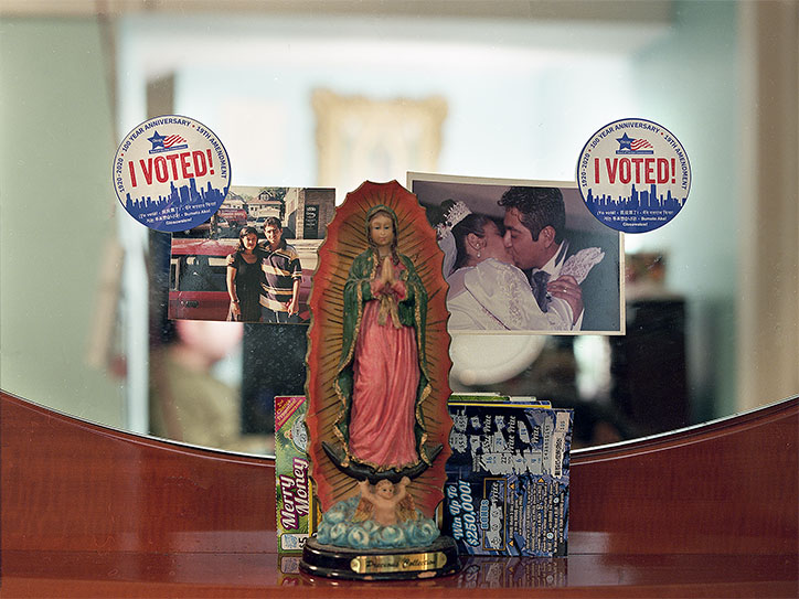 A close-up photograph of a dresser top with a mirror in the background. Centered in the image is a small statue of the Virgin of Guadalupe, placed in front of the mirror. Surrounding the statue are several personal items: on the left, there’s a photo of a young Mexican couple with their arms around each other; on the right, a photo shows the same couple as bride and groom sharing a kiss. Above each photograph is an "I VOTED!" sticker with a blue skyline design, celebrating the 30th anniversary of voting rights for certain communities. Behind the Virgin of Guadalupe statue, scratch-off lottery tickets and a small box labeled "Merry Money" are stacked. The setting reflects a blend of cultural identity, family values, and everyday life.